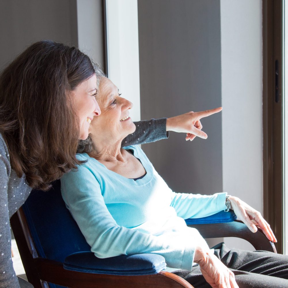 Positive mother and daughter enjoying dramatic view out of window. Young health visitor showing to senior lady funny scene out of window. Family relations concept
